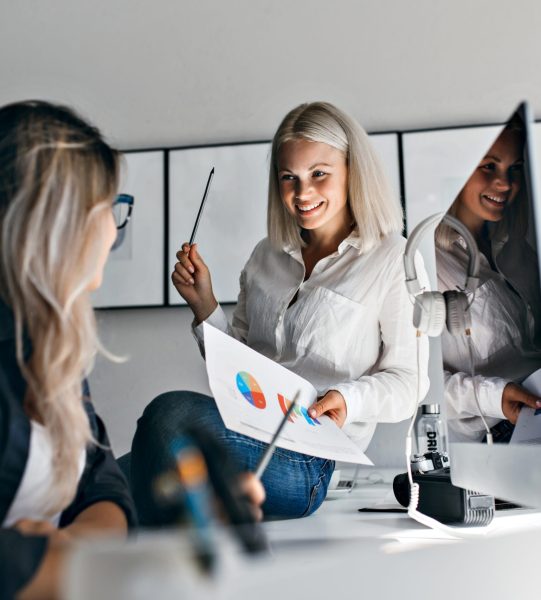 Smiling blonde female manager holding infographic and pencil, while sitting on table. Indoor portrait of two girls working with computer in office.